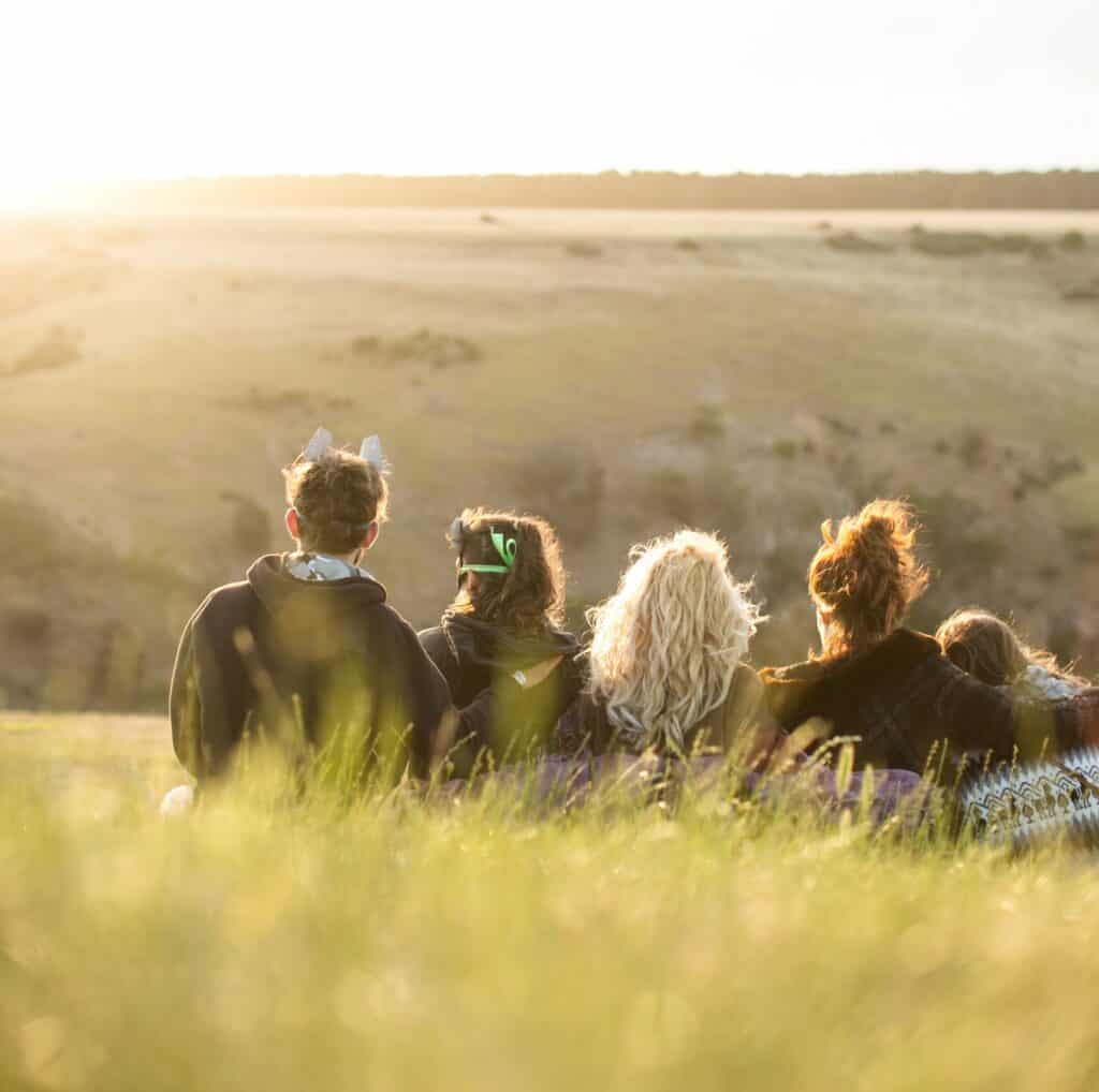 Five young people sit on a grassy hill watching the sun set over fields.